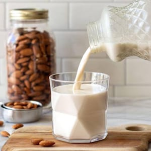 Puede incluir: Un vaso de leche de almendras que se vierte de una botella de vidrio decorativa. Un tarro de almendras está en el fondo, junto con una taza medidora de almendras y una tabla de cortar de madera. La leche de almendras es de color blanco cremoso.
