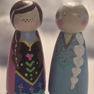 May include: Two hand-painted wooden peg dolls, one with brown hair and a pink and blue dress, the other with white hair and a blue dress. The dolls have painted faces with rosy cheeks and are standing upright.
