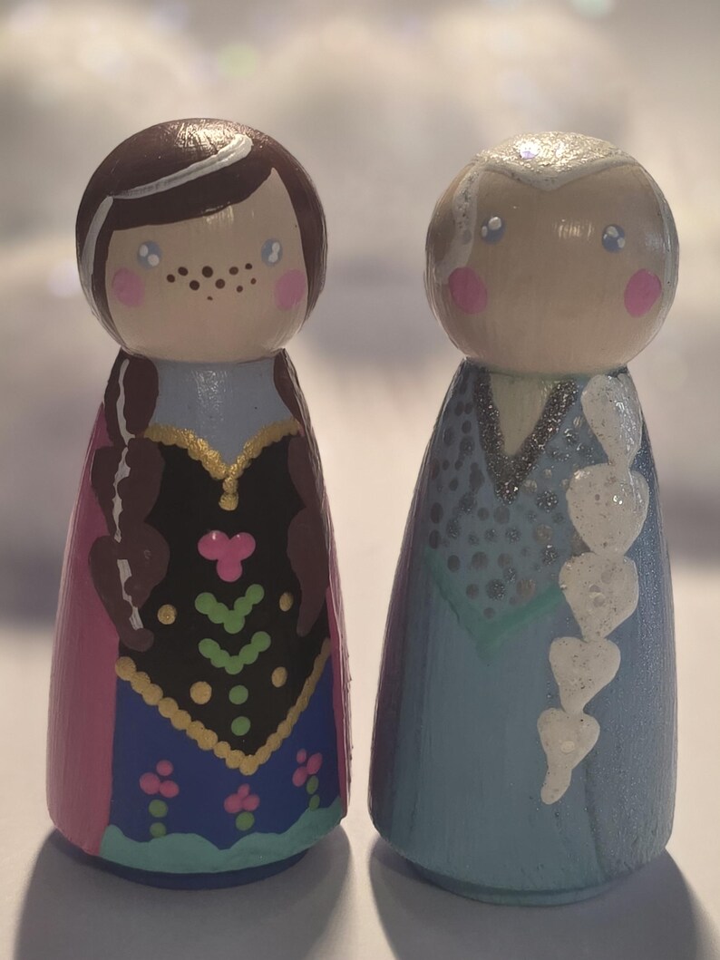 May include: Two hand-painted wooden peg dolls, one with brown hair and a pink and blue dress, the other with white hair and a blue dress. The dolls have painted faces with rosy cheeks and are standing upright.