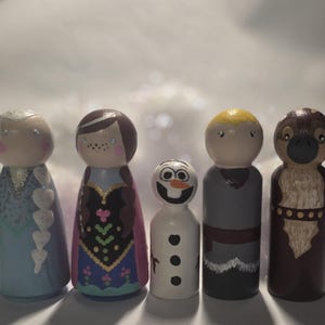 May include: Five wooden peg dolls painted to resemble characters from the animated film "Frozen." The dolls include Elsa, Anna, Olaf, Kristoff, and Sven. Each doll is hand-painted with detailed features and vibrant colors, standing approximately 4 inches tall.