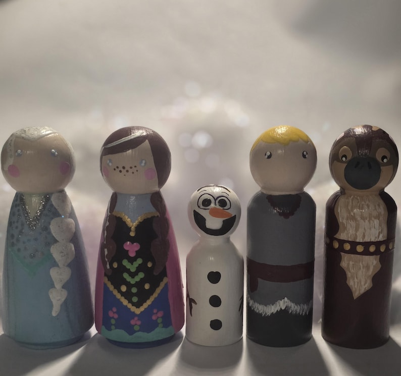 May include: Five wooden peg dolls painted to resemble characters from the animated film "Frozen." The dolls include Elsa, Anna, Olaf, Kristoff, and Sven. Each doll is hand-painted with detailed features and vibrant colors, standing approximately 4 inches tall.