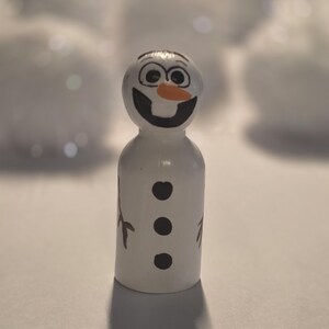 May include: A white wooden snowman figurine with a smiling face, black eyes, and an orange carrot nose. The snowman has three black buttons and brown twig arms. The figurine is standing upright.