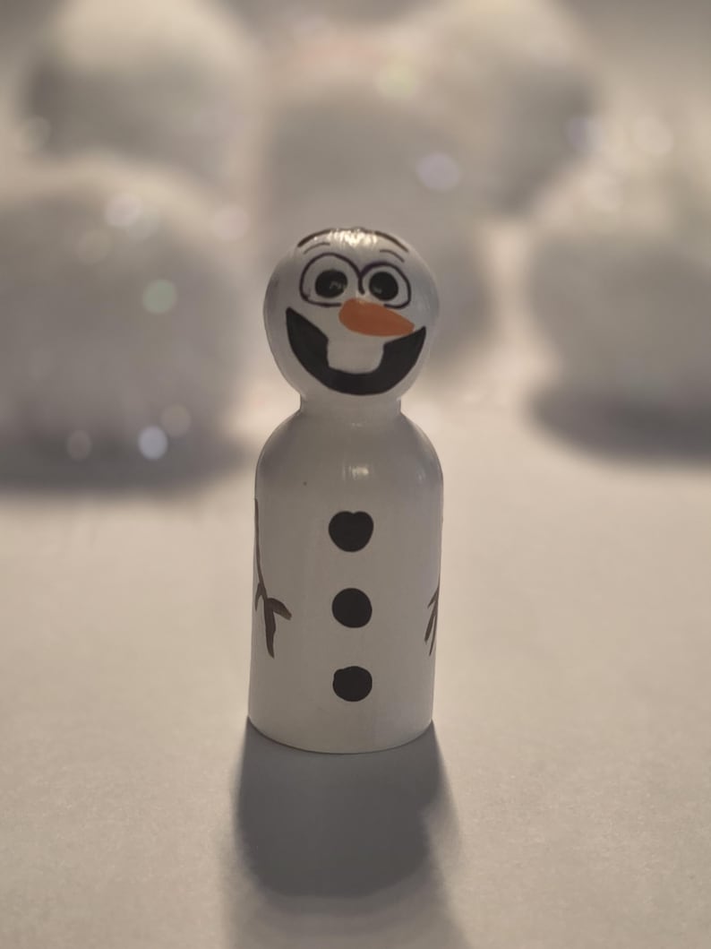 May include: A white wooden snowman figurine with a smiling face, black eyes, and an orange carrot nose. The snowman has three black buttons and brown twig arms. The figurine is standing upright.