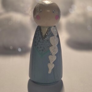May include: A wooden peg doll painted to resemble a character with a blue dress, white hair, and pink cheeks. The doll has a glittery finish and a white heart-shaped braid. The background is blurred with white shapes.