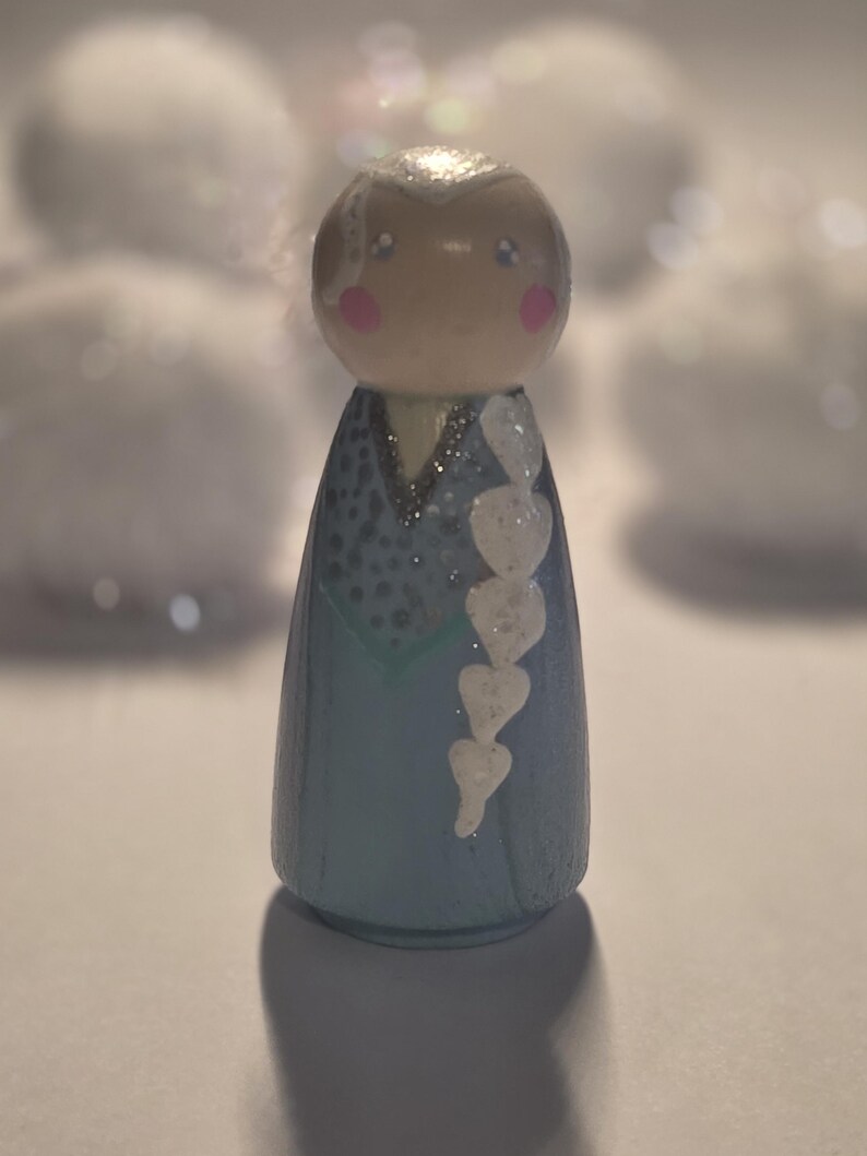 May include: A wooden peg doll painted to resemble a character with a blue dress, white hair, and pink cheeks. The doll has a glittery finish and a white heart-shaped braid. The background is blurred with white shapes.