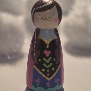 May include: A hand-painted wooden peg doll featuring a character with brown hair in braids, freckles, and rosy cheeks. The doll wears a blue and black dress with pink accents and a pink cape. The doll is approximately 4 inches tall.