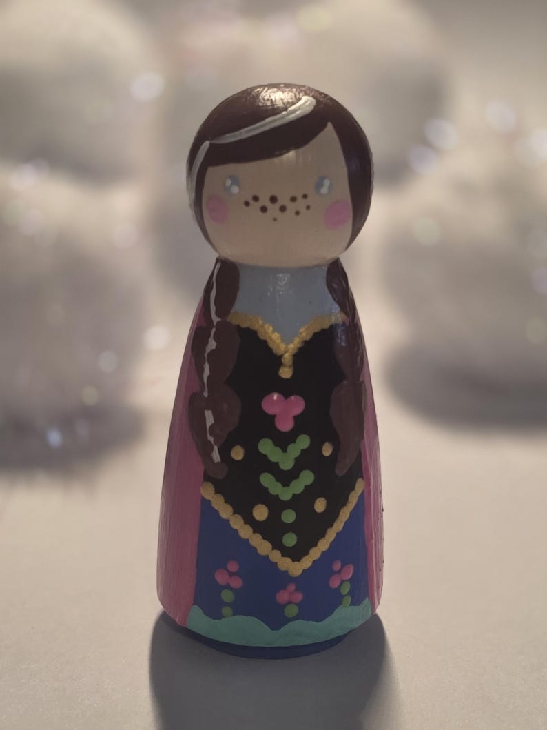 May include: A hand-painted wooden peg doll featuring a character with brown hair in braids, freckles, and rosy cheeks. The doll wears a blue and black dress with pink accents and a pink cape. The doll is approximately 4 inches tall.