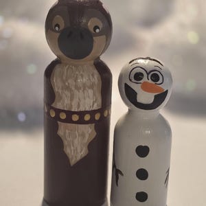 May include: Two wooden peg dolls, one resembling a cartoon owl with brown and tan paint, and the other a snowman with a carrot nose and black details. The owl doll has a dark brown body and a tan chest.