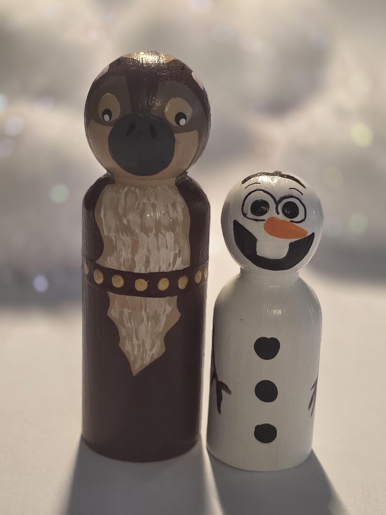 May include: Two wooden peg dolls, one resembling a cartoon owl with brown and tan paint, and the other a snowman with a carrot nose and black details. The owl doll has a dark brown body and a tan chest.