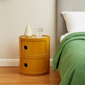 Puede incluir: Una mesita de noche de color amarillo mostaza con dos cajones y una parte superior redonda. Un pequeño jarrón blanco y un vaso de agua están encima. La mesa está junto a una cama con una manta verde y una almohada blanca.