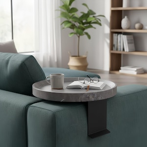Könnte beinhalten: Ein runder Beistelltisch in Grau mit Marmoroptik und einem schwarzen Metallstützarm ist an einem teal-farbenen Sofa befestigt. Eine Tasse, ein offenes Buch und eine Brille liegen auf dem Tisch. Eine Topfpflanze und ein Bücherregal befinden sich im Hintergrund.