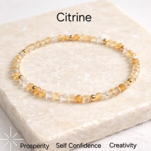 Könnte beinhalten: Ein zartes Armband aus kleinen, facettierten Citrin-Perlen in Gelb- und Klartönen. Das Armband ist mit kleinen goldfarbenen Perlen akzentuiert. Das Wort "Citrin" ist über dem Armband aufgedruckt. Die Wörter "Wohlstand", "Selbstvertrauen" und "Kreativität" sind darunter aufgedruckt.