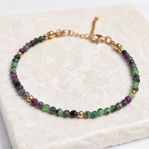 May include: A delicate beaded bracelet featuring small, faceted beads in shades of green, purple, and black, interspersed with gold-coloured metal beads. The bracelet has a gold-coloured chain and clasp closure.