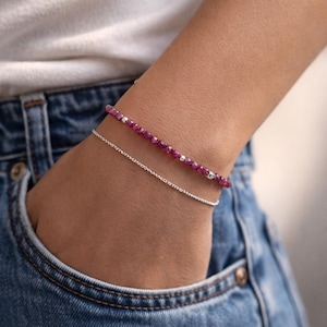 May include: Two bracelets are shown on a wrist. The top bracelet features small, faceted, deep pink beads with silver accents. The bottom bracelet is a delicate silver chain. The person is wearing a white top and blue denim jeans.