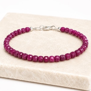 May include: A bracelet composed of small, faceted, deep magenta-coloured beads arranged in a continuous circular pattern. The piece features a silver-toned metal lobster claw clasp mechanism at the top, connecting the two ends of the beaded strand.