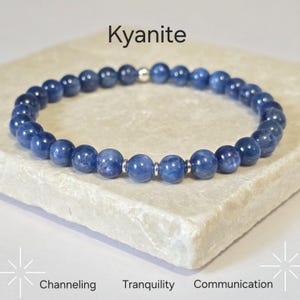 May include: A blue kyanite beaded bracelet with silver detailing. The bracelet is presented on a light stone surface. The image includes the text "Kyanite", "Channeling", "Tranquility", and "Communication".