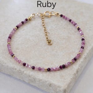 May include: A delicate bracelet with small, faceted pink and deep red gemstones and gold-coloured beads. The bracelet has an adjustable gold-coloured chain and clasp. The word "Ruby" is displayed in black text at the top.