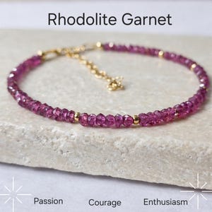 May include: A delicate bracelet featuring rhodolite garnet beads in shades of pink and purple. The bracelet is strung on a gold chain with gold accents. Text on the image reads "Rhodolite Garnet", "Passion", "Courage", and "Enthusiasm".
