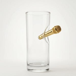 Può includere: Un bicchiere trasparente di forma cilindrica, attraversato da un microfono dorato. Il microfono presenta una testa strutturata e un corpo liscio. Il tutto è posto su uno sfondo bianco, evidenziando il design originale.