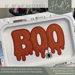 May include: An embroidery hoop containing white fabric with the word BOO stitched in orange glitter thread. The letters have a dripping effect, and a small black spider is embroidered below the second O. The hoop frame has warning labels.