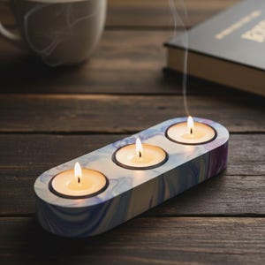 May include: A decorative candle holder with three lit candles. The holder has a marbled blue, purple, and white design. Smoke rises from the candles. A white mug and a book are in the background on a wooden surface.