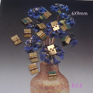 Può includere: Primo piano di 14 cerniere in metallo dorato, ciascuna misura 6x9 mm. Le cerniere sono sparse su una superficie nera con un vaso di fiori blu sullo sfondo. Il testo "6X9mm" è visibile nell'angolo in alto a destra dell'immagine.