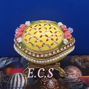 Puede incluir: Un huevo de Pascua amarillo, tallado y decorado de forma intrincada, con un borde dorado y detalles de perlas. El huevo está adornado con flores rosas y azules y se asienta sobre un soporte dorado. El texto "E.C.S." es visible en la imagen.