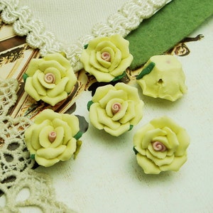 May include: Six pale yellow ceramic rose flower embellishments with pink centers. The roses are arranged on a white surface with a lace trim.