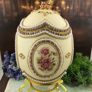May include: A white egg-shaped decorative box with a gold stand and a pink floral design. The box is decorated with pearls and rhinestones.