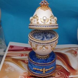 Può includere: Una scatola musicale a forma di uovo in porcellana bianca con dettagli dorati e blu e bianchi. La scatola è decorata con perle e strass. Il coperchio è aperto, rivelando un interno in velluto blu. La scatola è appoggiata su una base in velluto blu con finiture dorate.