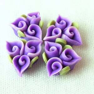 Puede incluir: Cinco adornos florales de arcilla polimérica de color morado con hojas verdes. Cada flor está hecha de tres formas de flores individuales con un remolino en el centro.