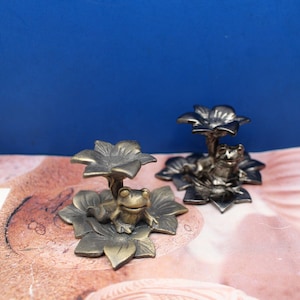 May include: Two frog figurines sitting on lily pads. The frog on the left is gold, and the frog on the right is silver.