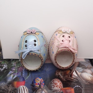 May include: Two intricately decorated Easter eggs, one blue and one pink, are shaped like shoes with laces and floral accents. The eggs are sitting on a surface with other decorated eggs.