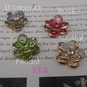 Könnte beinhalten: Vier blütenförmige Verzierungen mit goldenen Fassungen und verschiedenen farbigen Glassteinen. Die Steine sind hellblau, rosa, grün und klar. Der Text "Lt Sapphire", "ROSE", "Crystal" und "Peridot" ist auf dem Bild zu sehen. Der Text "E.C.S" ist ebenfalls auf dem Bild zu sehen.