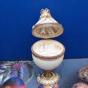 May include: An open, decorative egg-shaped container with a white exterior and gold trim. The interior is lined with a satin-like fabric. The top is adorned with a gold crown-like embellishment. The egg sits on a decorative stand.