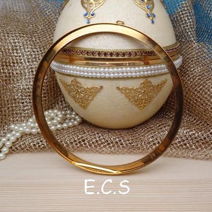 Peut inclure: Un bracelet jonc doré est au premier plan, avec un récipient décoratif en forme d'œuf derrière lui. L'œuf est de couleur crème avec des accents dorés et de bijoux, et un rang de perles est drapé à proximité. Les lettres "E.C.S" sont en bas.
