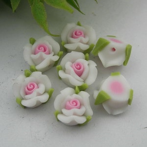 Puede incluir: Una colección de cuentas de cerámica blanca en forma de rosa con centros rosados y hojas verdes. Las cuentas son perfectas para la elaboración de joyas, el scrapbooking y otros proyectos de manualidades.