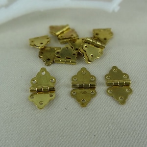 Puede incluir: Un grupo de 10 bisagras de metal dorado con un diseño decorativo. Las bisagras son pequeñas y tienen un aspecto delicado y ornamentado.