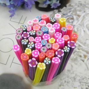 Puede incluir: Una colección de palitos de arte de uñas de flores de arcilla polimérica de colores. Los palitos están dispuestos en un recipiente con forma de jarrón y presentan una variedad de diseños de flores, incluyendo rosas, moradas, amarillas y blancas.
