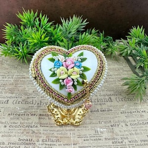 May include: Heart-shaped trinket box with a floral design. The box features a white interior with a bouquet of pink, blue, and yellow flowers. The exterior is trimmed with pearls, pink gems, and gold accents. The box sits on a decorative gold base.