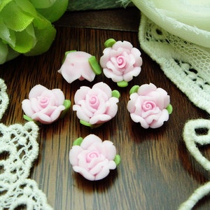 Può includere: Primo piano di sei piccoli abbellimenti a forma di rosa rosa chiaro con foglie verdi. Le rose sono disposte su una superficie di legno scuro, con pizzo e verde sullo sfondo. Questi elementi decorativi sono ideali per progetti di artigianato.