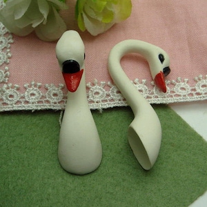 May include: Two white ceramic swan figurines with red beaks. The swan on the left is standing upright, while the swan on the right is curved into a spoon shape.