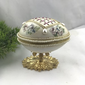 May include: An egg-shaped decorative box with a floral design and pearl accents. The off-white box features a gold base and is adorned with pink gemstones. The egg-shaped box is a decorative item.