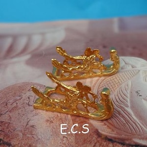 May include: Two gold-colored metal sword stands with intricate floral designs. The stands are shaped like a pair of stylized sleighs.