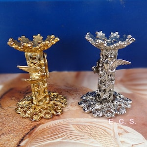 Puede incluir: Dos soportes decorativos, uno dorado y otro plateado, con diseños intrincados que presentan un pájaro con las alas extendidas. Los soportes tienen una parte superior redonda y una base con un patrón floral.