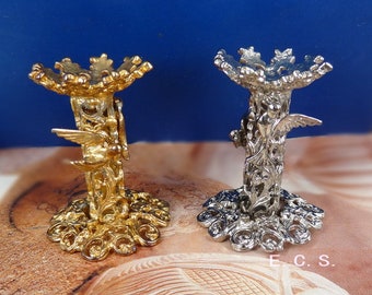 May include: Two decorative stands, one gold and one silver, with intricate designs featuring a bird with wings outstretched. The stands have a round top and a base with a floral pattern.