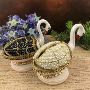 Op de afbeelding: Twee witte zwanen-vormige muziekdozen met bejeweled deksels. Een deksel is versierd met zwarte en gouden kralen, de andere met witte parels.
