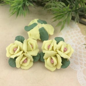 May include: Close-up of three handmade yellow rose flower embellishments with green leaves and pink centers. The flowers are arranged on a light-colored surface, with green foliage in the background. These decorative flowers are ideal for craft projects.