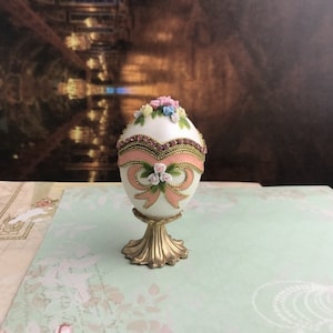 May include: A white porcelain egg with a pink ribbon and floral accents, sitting on a gold stand. The egg is decorated with rhinestones and has a delicate floral design.