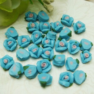 May include: A collection of small, blue, ceramic rose flower embellishments with pink centers. The roses are scattered on a white surface.